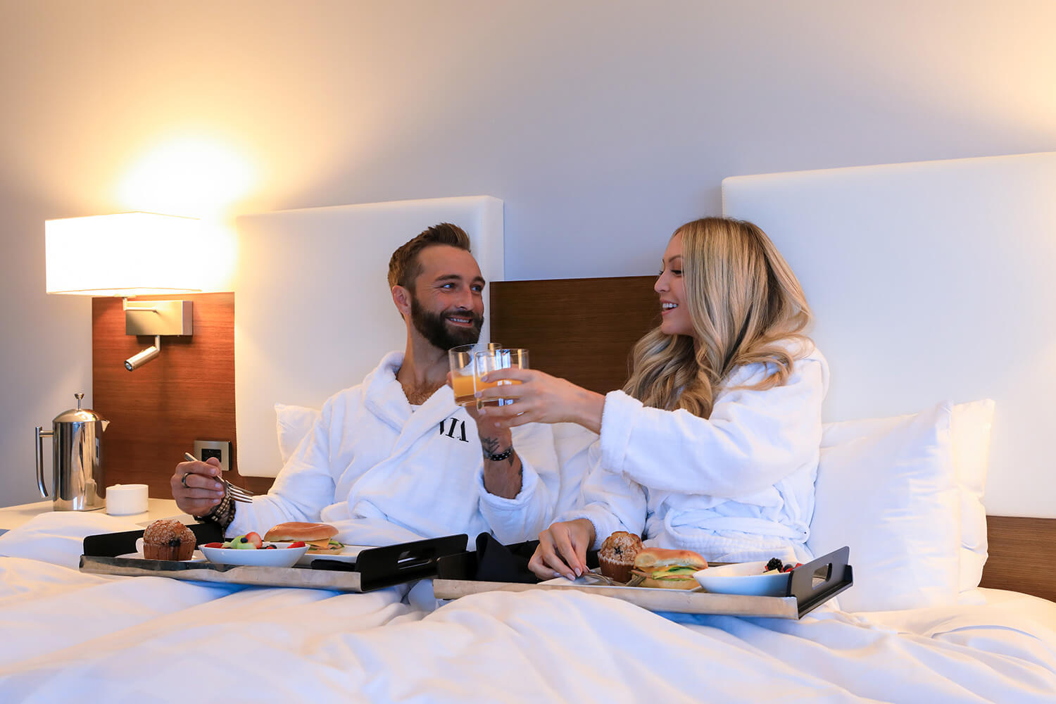 A man and woman in bathrobes share a Valentine's meal in bed, holding trays with various foods and drinks. They appear to be toasting with glasses, sitting under white bedding in a well-lit room.
